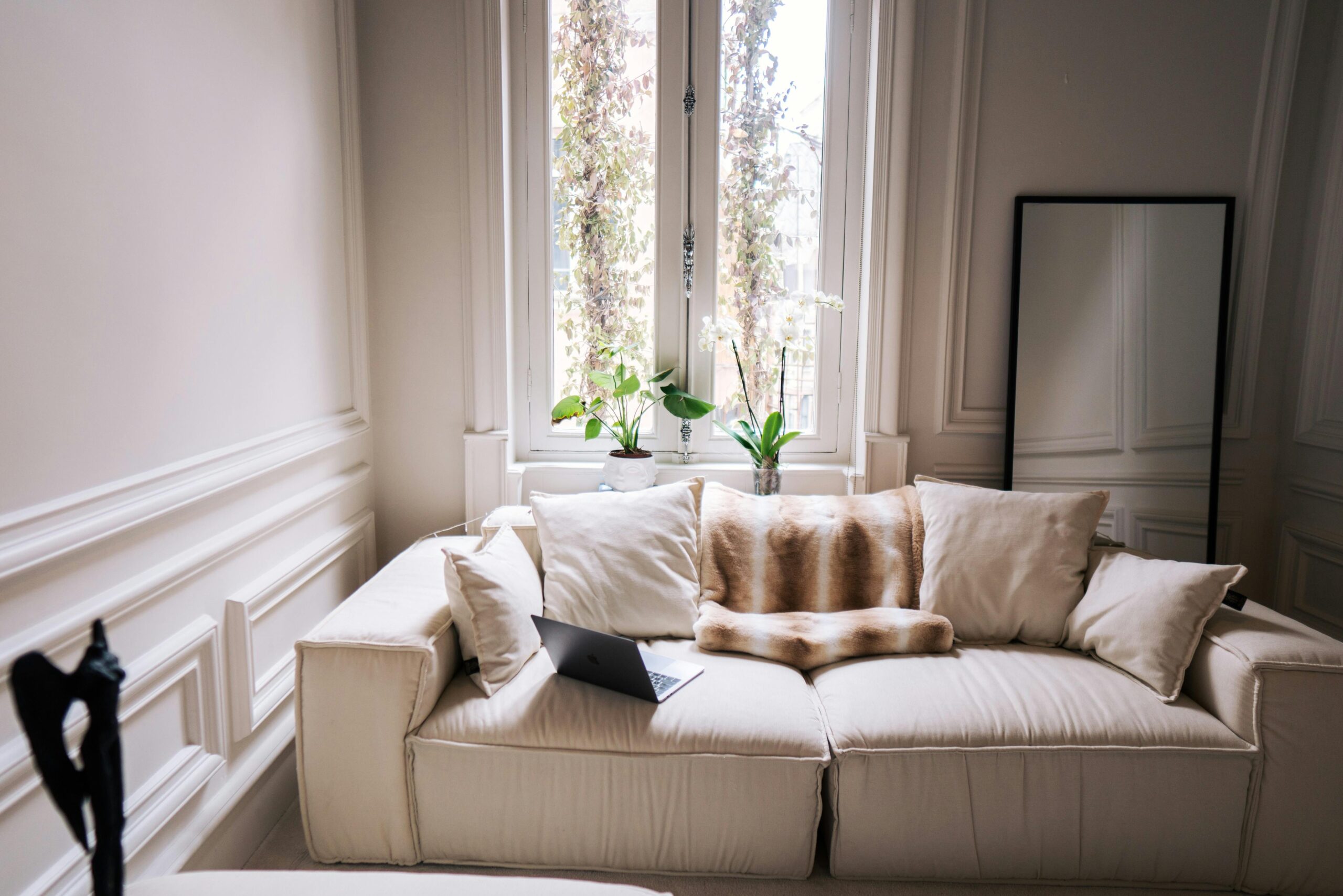 A modern living room featuring a comfortable beige sofa, indoor plants, and a large window for natural light.